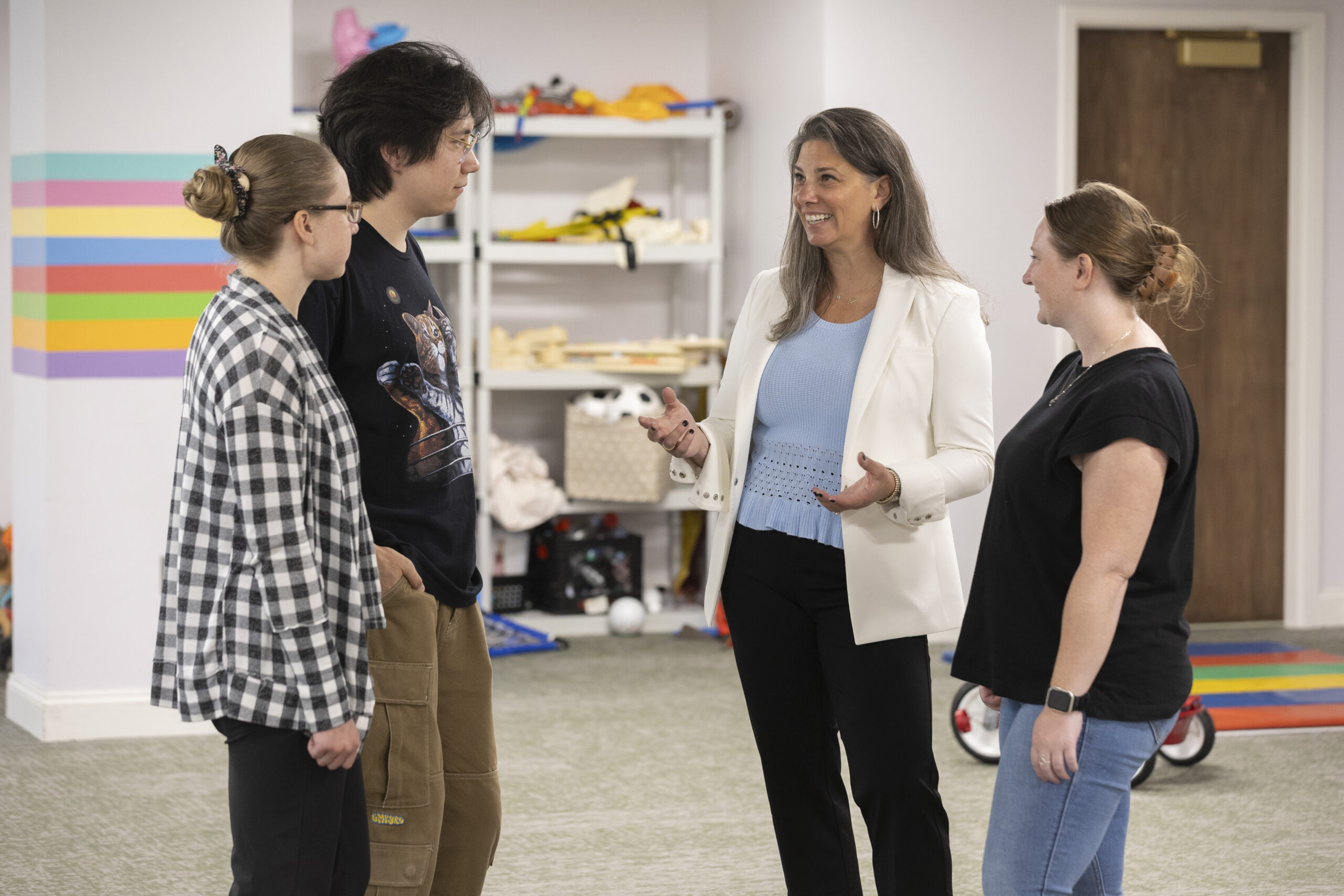 A group of four caucasian adults are standing and talking as a group, while there are toys and colorful mats and walls in the background.