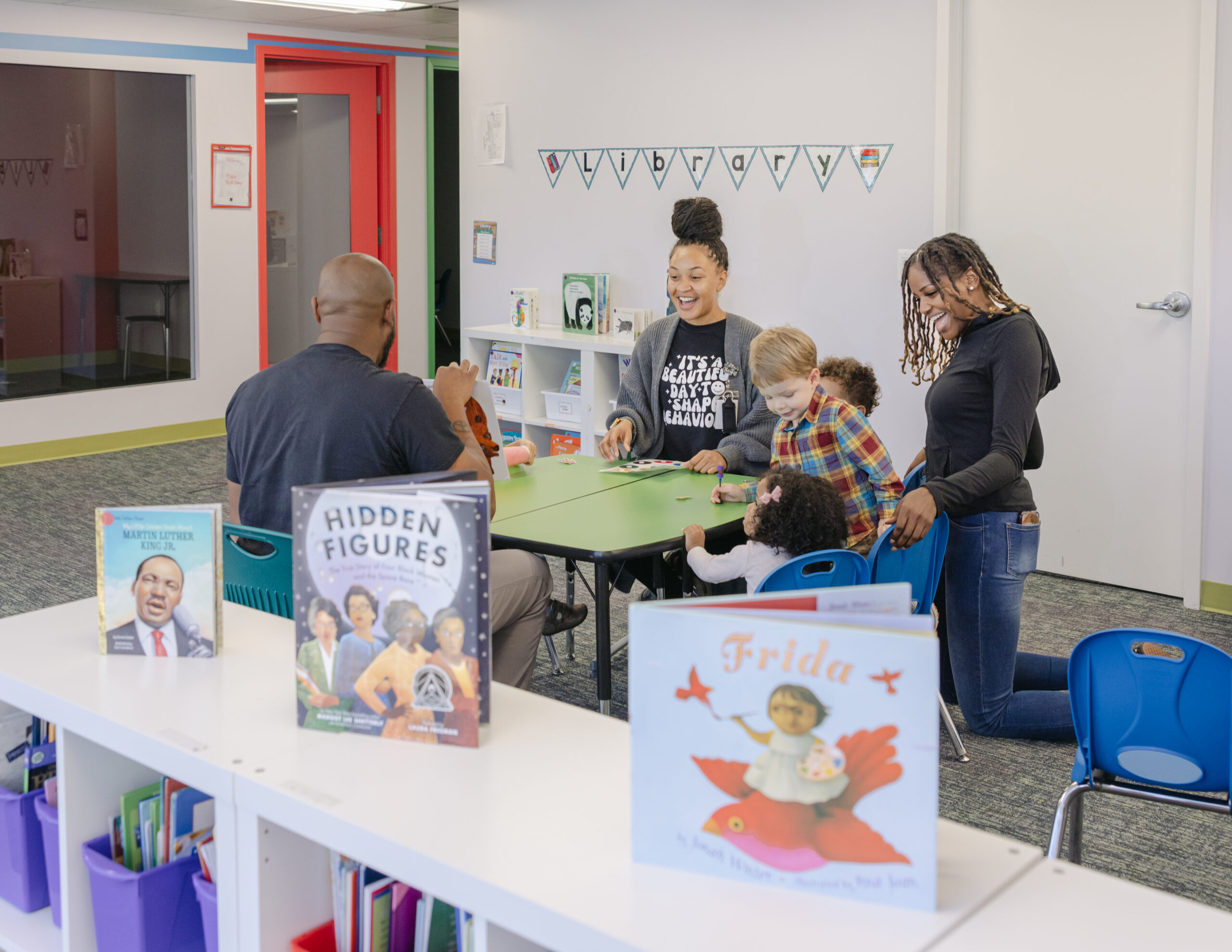 A group of african-american adults and caucasian children sit around a green table in a therapy office. Books are displayed on a white shelf in the foreground.