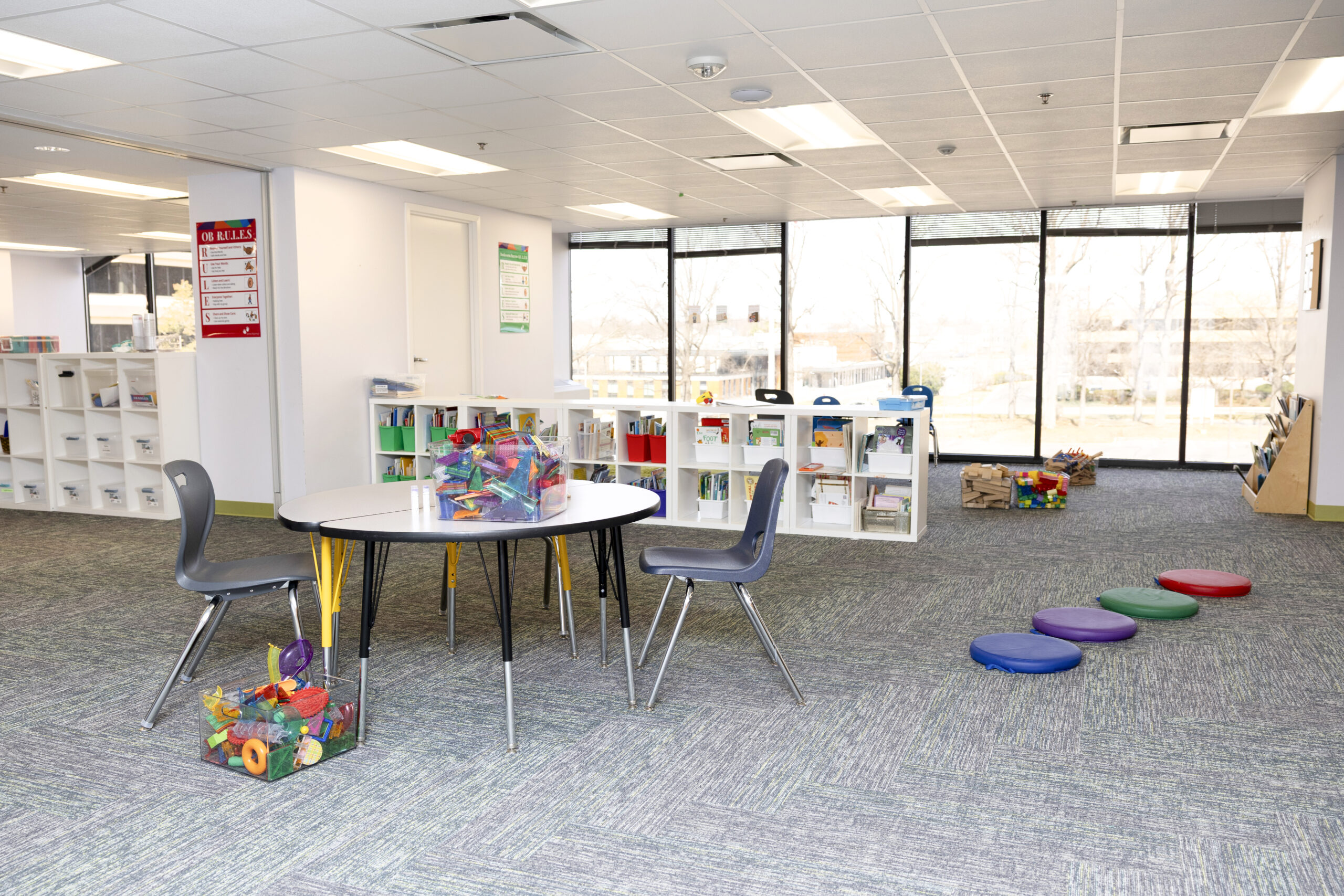 A picture of an empty classroom with white storage cubbies, a table with two chairs and a bin of toys on top, along with other colorful toys around the room.